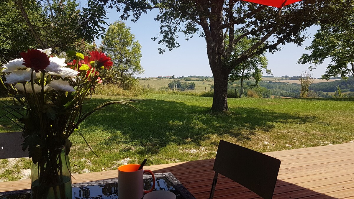 A sunlit wooden terrace is shown with a vase of flowers and a coffee cup on a table. The expansive garden is visible, featuring lush green grass and distant rolling hills, under a clear blue sky and shaded by a large tree.