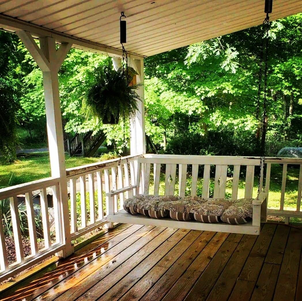 A welcoming porch features a suspended swing adorned with a cushioned seat, surrounded by lush greenery. Sunlight filters through the trees, illuminating the wooden decking and creating a serene outdoor seating area.