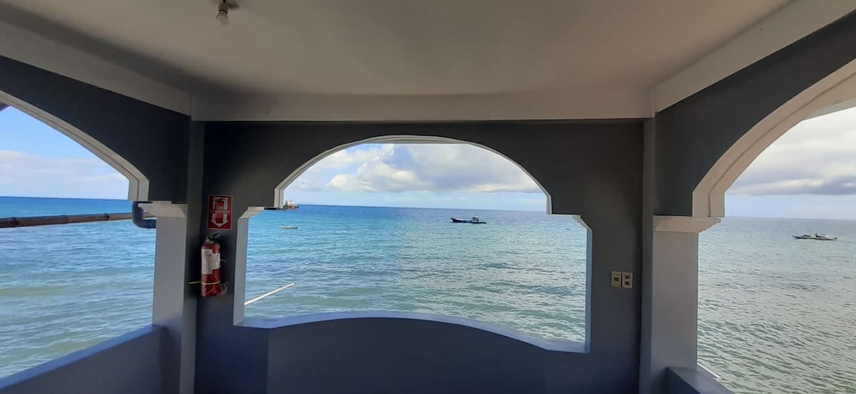 A spacious archway offers a clear view of the calm sea, where small boats can be seen on the horizon. The scene is framed by a light gray wall, enhancing the open and airy feel, with soft cloud formations visible in the sky.