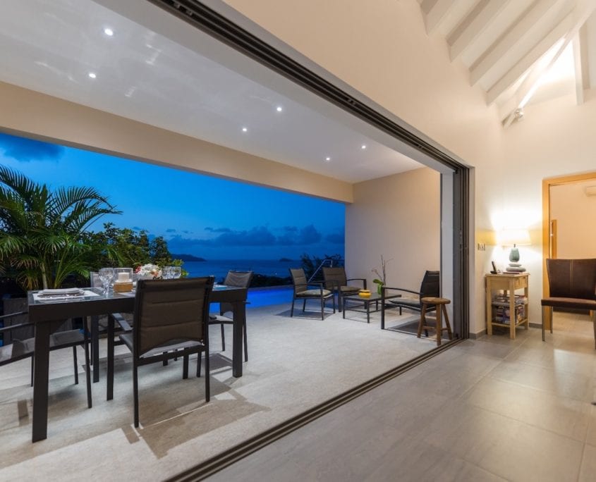 An open-concept living area features large sliding glass doors revealing a panoramic view of the bay at dusk. A dining table with black chairs sits comfortably on the terrace, while a cozy seating area is positioned nearby with warm light from a table lamp.