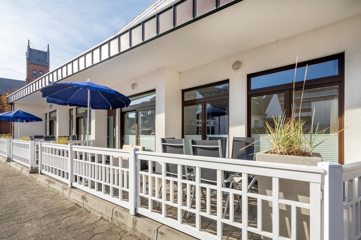 A spacious outdoor seating area is featured, with a row of tables and chairs set against a backdrop of large windows. Two blue umbrellas provide shade, while potted plants add a natural touch. The structure's design incorporates elements of modern architecture.