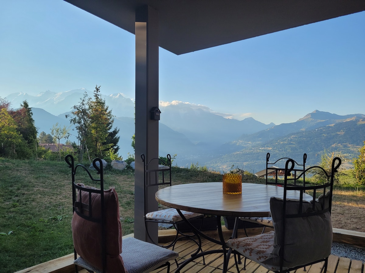 A covered terrace features a round wooden table surrounded by four wrought-iron chairs. Behind, panoramic views of distant mountains and valleys are visible, framed by trees and greenery. Soft sunlight enhances the scenic backdrop, creating a peaceful outdoor dining space.