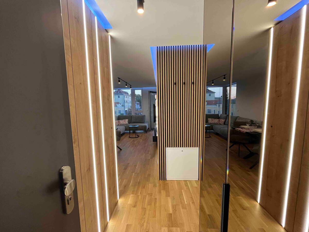 The entrance features a modern hallway with sleek wooden paneling and illuminated accents. Natural light filters through large windows in the background, revealing an open living space furnished with comfortable seating and contemporary decor.