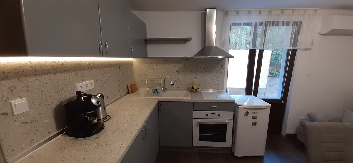 A modern kitchen is equipped with sleek cabinetry and a textured countertop. Essential appliances include a coffee maker, oven, and refrigerator. Natural light enters through large windows, complementing the neutral color scheme. A cozy seating area is visible in the background.