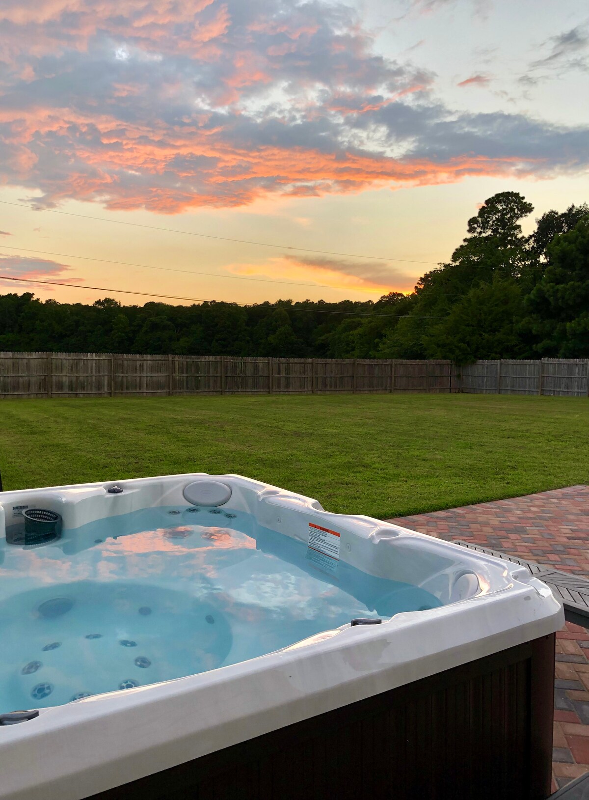 A newly added hot tub is depicted on the patio, with clear blue water reflecting the vibrant colors of the sunset. The spacious, grassy backyard is enclosed by a wooden fence, offering a serene setting amidst the natural surroundings.