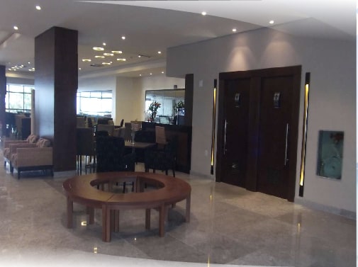 A modern lobby area features a circular wooden coffee table at its center, surrounded by upholstered seating. Bright overhead lighting enhances the space, while large windows allow natural light to flow in, creating a welcoming environment for guests.