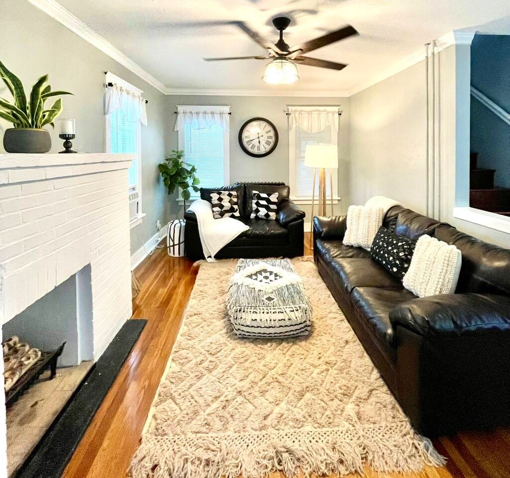 A living room features a leather sofa and a decorative armchair, complemented by a patterned area rug. A round clock hangs on the wall, and natural light filters through the windows. A cozy plant adds a touch of greenery to the inviting space.