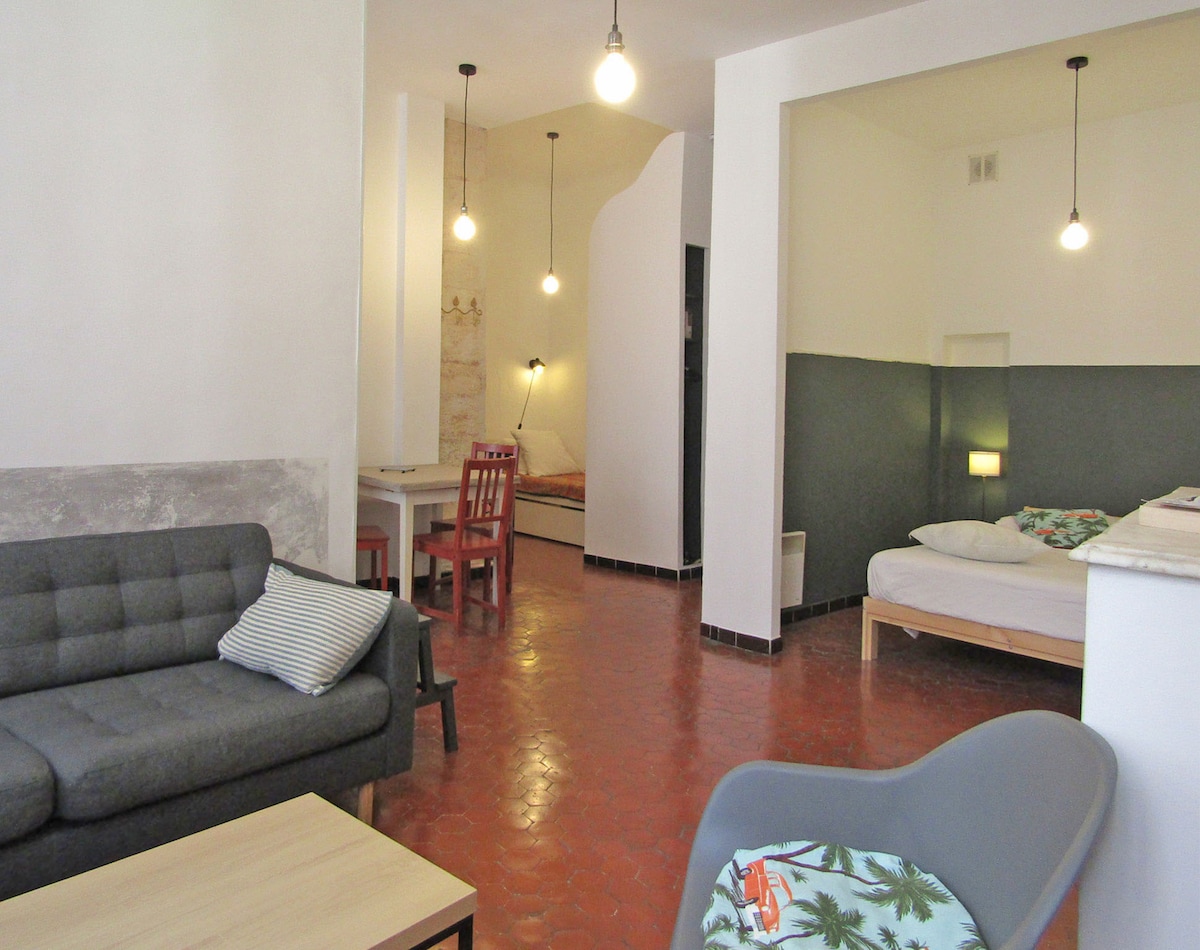 A welcoming living space features a comfortable gray sofa paired with a wooden coffee table. A dining area is visible, along with two sleeping alcoves equipped with double beds, each softly illuminated. The rustic tiled floor contrasts with the light walls, enhancing the room's spacious feel.