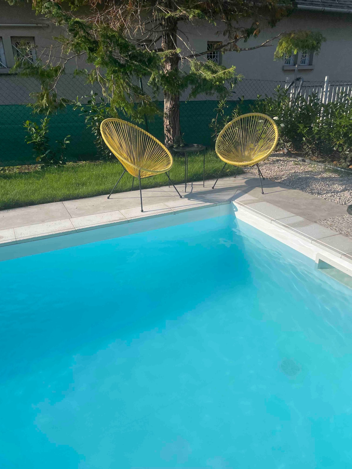 A tranquil pool area features clear blue water bordered by a sleek tiled edge. Two yellow chairs are positioned beside a small table on the pool deck, complemented by greenery in the background, creating an inviting space for relaxation.
