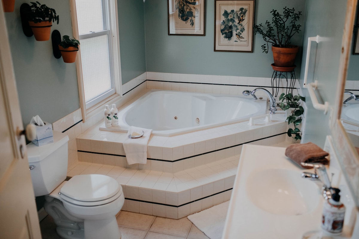 The spacious bathroom features a large Jacuzzi garden tub surrounded by white tile. A well-lit mirror is present above the sink, accented by fresh plants in wall pots. Natural light filters in through a nearby window, enhancing the calming atmosphere.