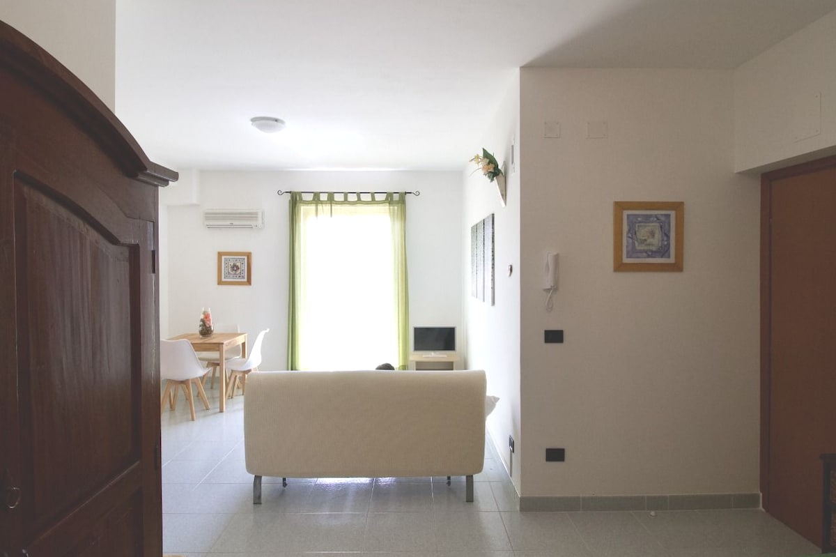 The interior space features a light-filled living area with a white sofa facing a small television. A dining table with chairs is visible in the background, complemented by a window with sheer green curtains. Wall art adds visual interest to the bright, minimalistic decor.