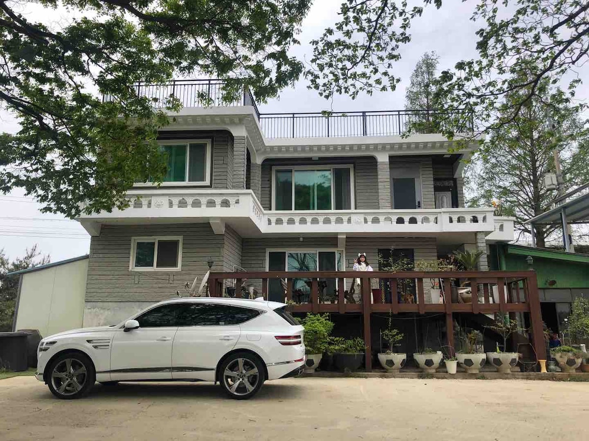 The two-story house features a balcony adorned with decorative railings and a spacious deck lined with potted plants. A sleek white vehicle is parked in front, complemented by lush trees in the surrounding area, enhancing the tranquil setting.