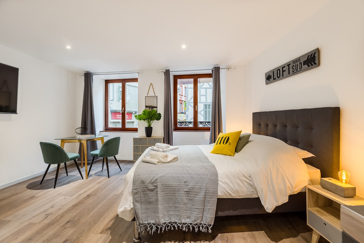 A comfortable sleeping area features a spacious bed with a light-colored blanket. Natural light filters through two windows, creating a bright atmosphere. A minimalist dining space is visible, including a small round table and two green chairs. Decorative plants and wall art add a touch of style.