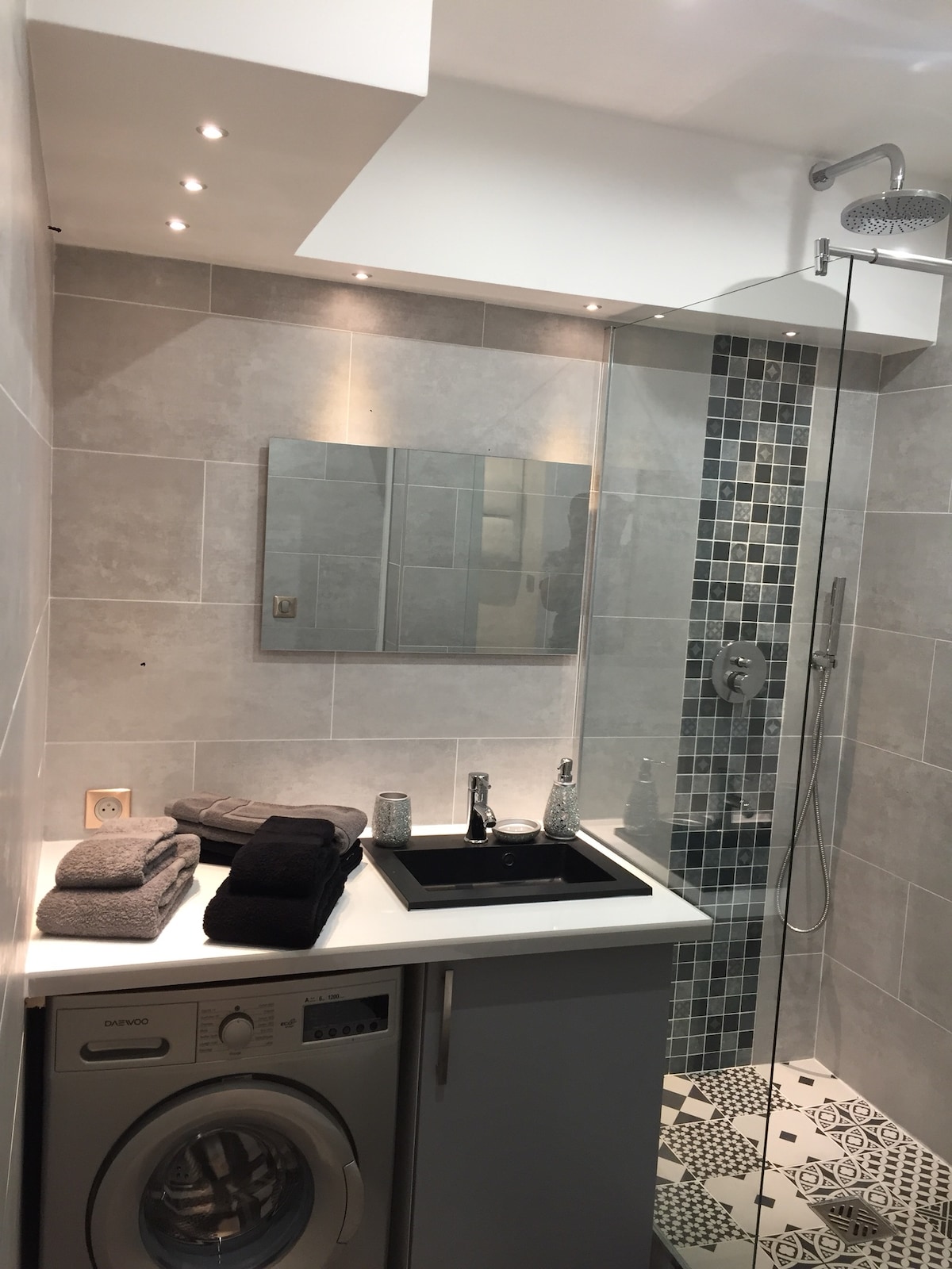 A modern bathroom features a walk-in shower with a glass enclosure and a stylistic wall of mosaic tiles. A compact washing machine is set beneath a sleek countertop, complemented by neatly arranged towels. Soft lighting enhances the contemporary design.