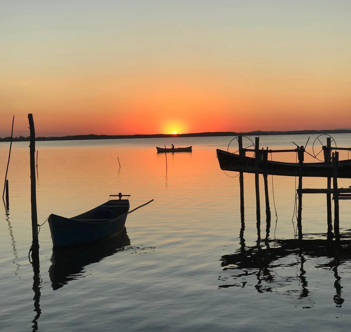 A serene sunset over the calm waters of the lagoon is captured, with silhouetted boats gently floating. Reflections of the vibrant orange and pink sky can be seen on the water's surface, creating a tranquil evening scene.