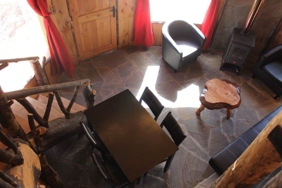 A cozy living area features a black dining table surrounded by four chairs. Natural wood elements and stone flooring enhance the rustic charm. Light streams through large windows covered with red curtains, creating a warm ambiance. A wood-burning stove adds functionality and warmth.