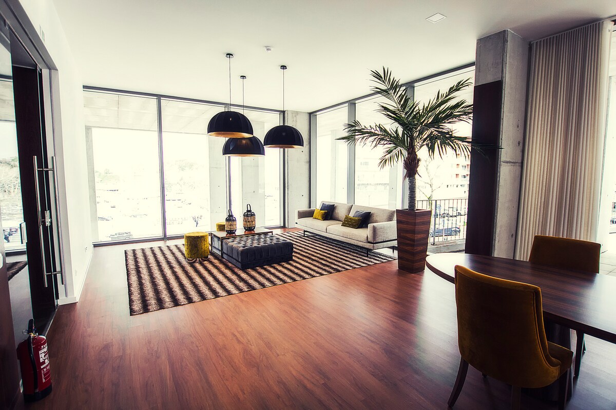 An open living area is illuminated by natural light through large windows. A cozy sofa and stylish coffee table are situated on a patterned rug, while two contemporary pendant lights hang overhead. A tall indoor plant adds a touch of greenery to the space.