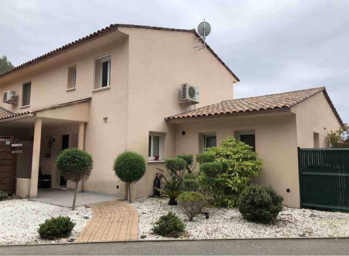 The exterior of a two-story house is presented, featuring a pink façade with terracotta roof tiles. Air conditioning units are visible, along with a landscaped front garden containing various shrubs and small hedges. A stone path leads to the entrance, enhancing accessibility.