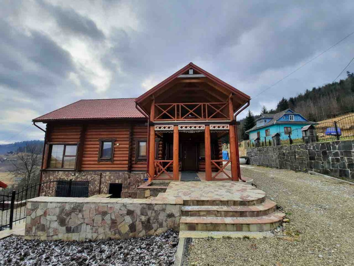 A charming wooden house with a sloped roof is set against a backdrop of rolling hills. The front terrace features a stone pathway and steps leading to the entrance. Surrounding greenery highlights the home's rustic appeal.