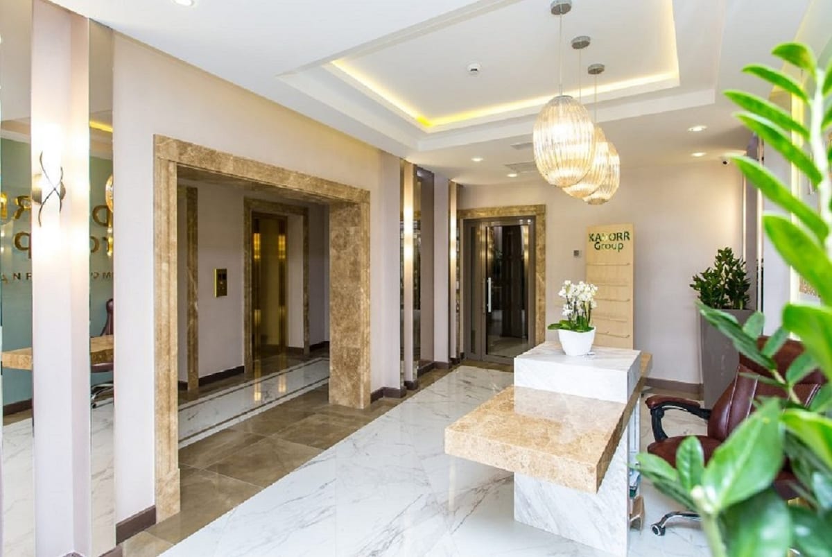 A modern lobby space is presented, featuring marble flooring and elegant lighting fixtures. A reception desk with a light-colored stone surface is positioned in front, complemented by green plants. Glass elevators are visible in the background, enhancing the inviting atmosphere of the entrance.