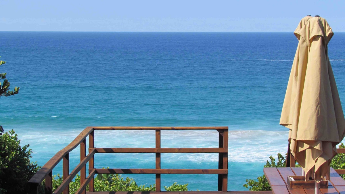A wooden deck offers a clear view of the expansive ocean, showcasing vibrant blue waters under a bright sky. A beige umbrella stands on one side, providing shade, while lush greenery frames the scene, enhancing the natural beauty of the coastal landscape.