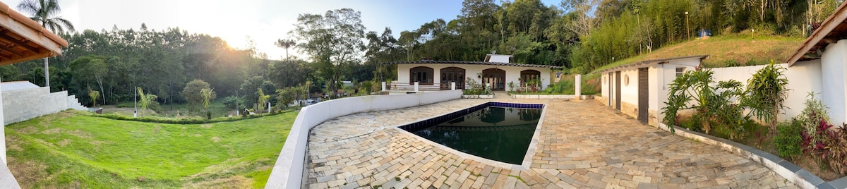 A panoramic view of a spacious outdoor area featuring a stone patio surrounding a rectangular pool. Lush greenery borders the property, while a single-story building is visible in the background, showcasing arched windows and a slanted roof that integrates with the natural setting.