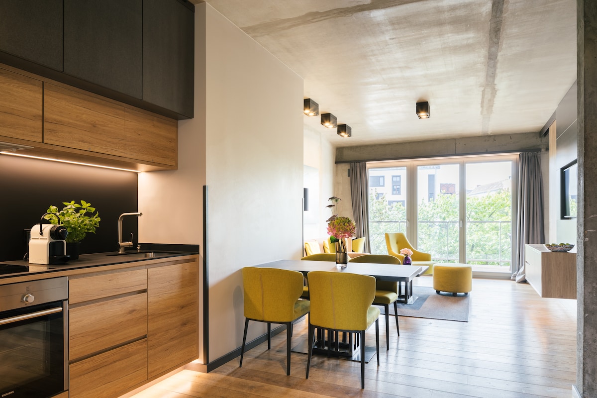 A modern kitchen area features wooden cabinetry and a sleek countertop, complemented by a coffee machine and a potted plant. Adjacent is a dining space with a table surrounded by yellow chairs. A spacious living area is visible, with large windows allowing natural light and offering views of greenery.