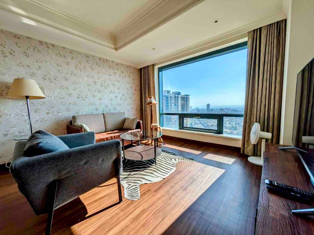 A spacious living area features large windows offering scenic city views. The room is furnished with a gray sofa, a brown armchair, and a round coffee table, complemented by a decorative rug. Natural light brightens the space, highlighting the floral wallpaper.