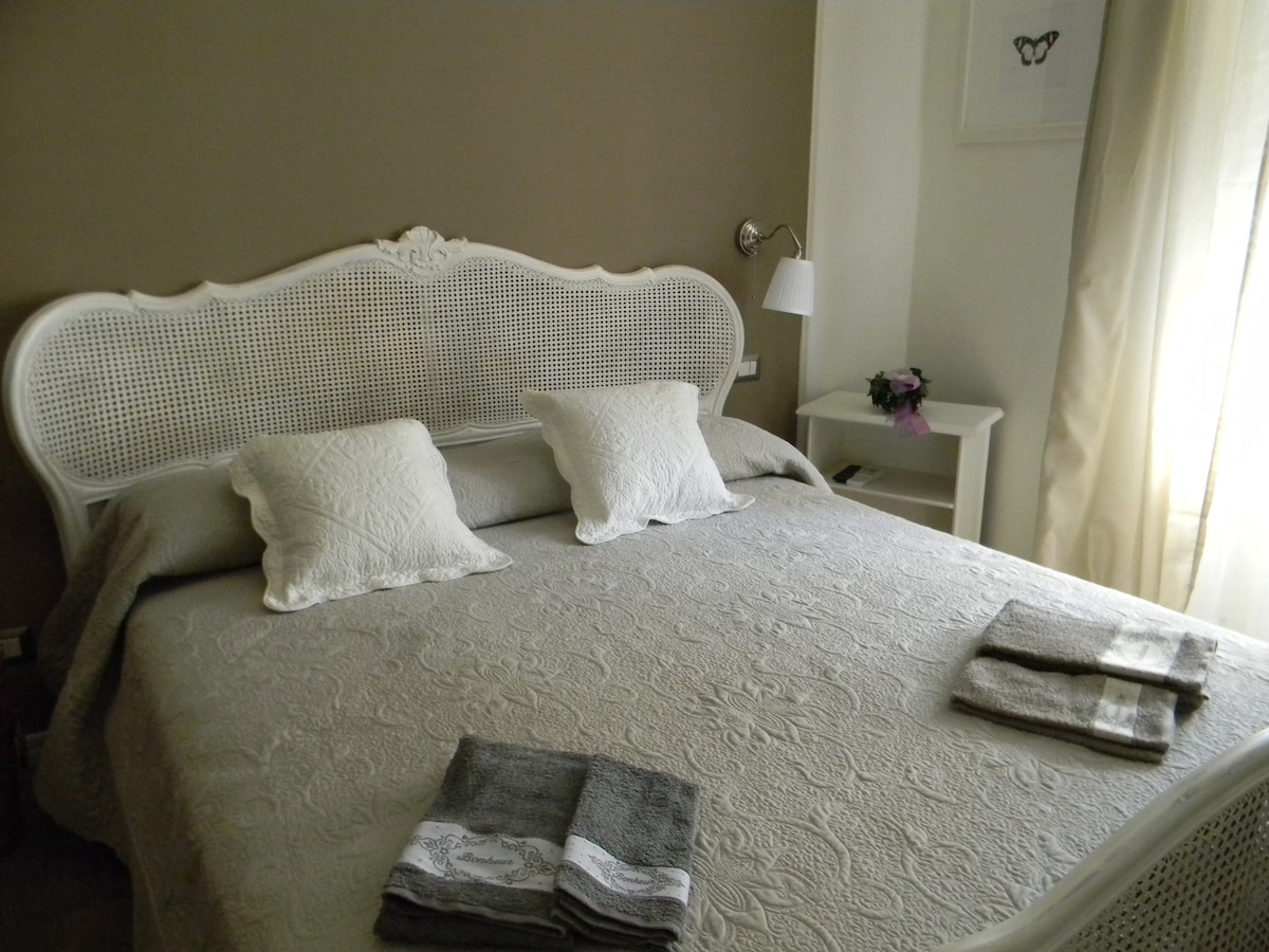 A spacious bed is dressed in a light quilt with textured patterns, complemented by two decorative pillows. Soft, neutral tones create a calm atmosphere, while natural light filters through sheer curtains. A bedside lamp and a small table enhance the room's functionality.