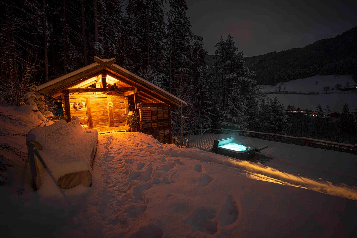 A rustic sauna cabin is illuminated warmly against a snowy backdrop, providing a contrast to the cold surroundings. Nearby, a hot tub is visible, with faint light highlighting its inviting water. The scene is framed by tall trees blanketed in snow.