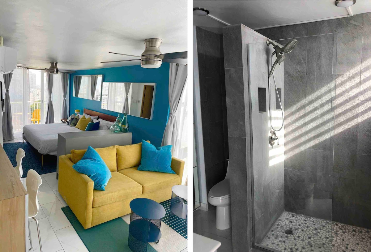 The open-concept studio presents a vibrant seating area with a yellow couch and blue accent pillows. A spacious bed is seen in the background, complemented by large windows that offer natural light. A neutral dining table and chairs complete the modern design.