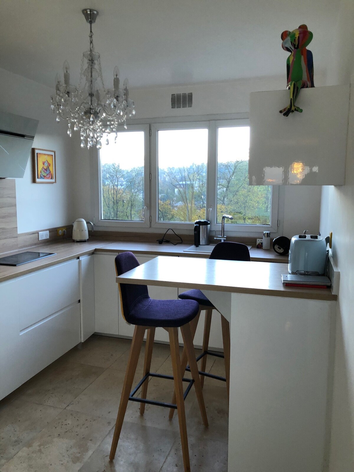 A modern kitchen features wooden countertops and a small breakfast bar with two high chairs. Natural light streams in through large windows, and a decorative chandelier hangs above. A colorful parrot figurine adds a playful touch to the sleek white cabinetry.