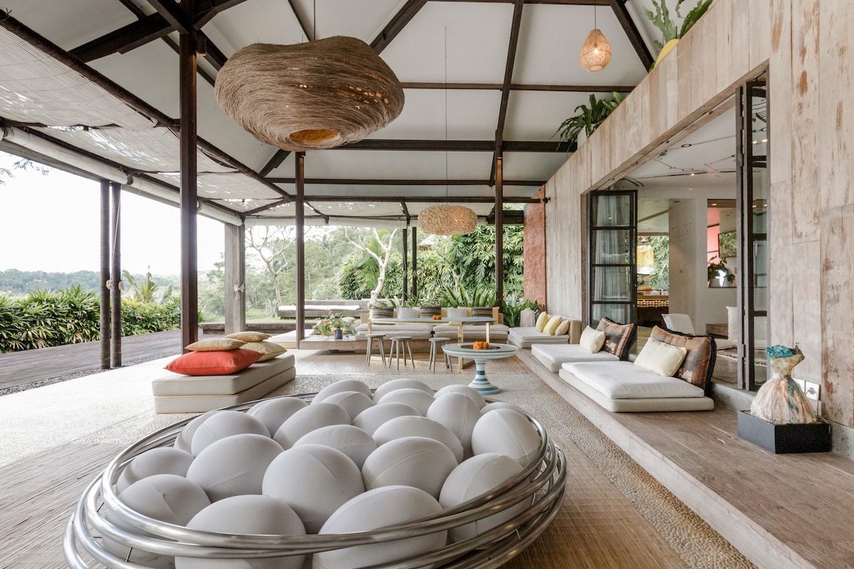 An expansive living area showcases a blend of natural materials, including polished concrete and wooden beams. Comfortable seating is arranged around a striking round sofa with white cushions. Large glass doors open to a picturesque outdoor space filled with greenery.