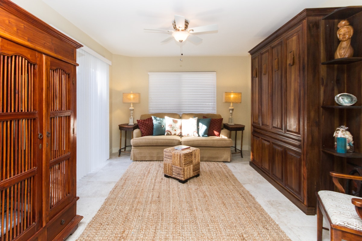 A spacious living area is prominent, featuring a comfortable couch adorned with decorative pillows. Two lamps offer soft illumination on either side. A textured rug lies on the floor, complemented by a natural fiber ottoman and wooden furniture against the walls.