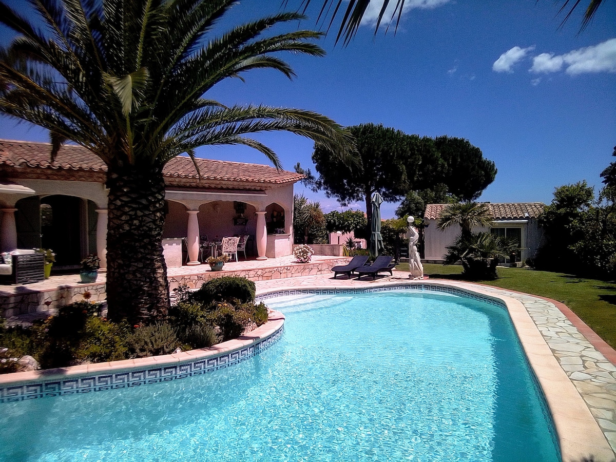 A serene pool area is surrounded by palm trees and lush greenery, with a clear blue water reflecting the sky. Sun loungers are positioned near the edge of the pool, offering a tranquil space to relax. The villa's exterior features rustic stonework and inviting outdoor seating.