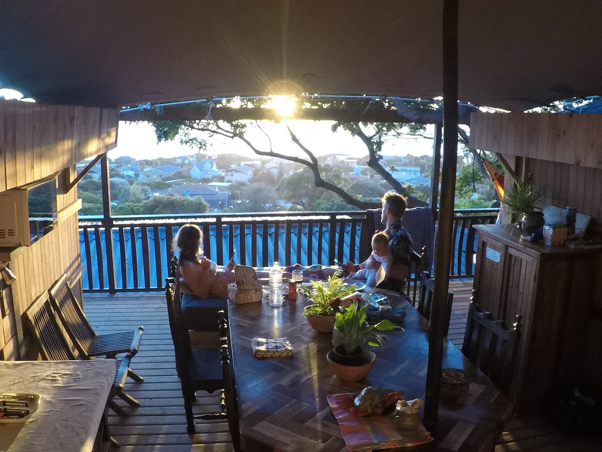 A spacious wooden deck is illuminated by the soft glow of sunset, with views of surrounding nature. A large table is set for dining, surrounded by several chairs. Potted plants are placed on the table, enhancing the outdoor dining experience.