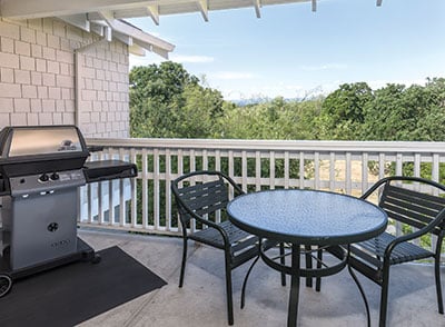 A private balcony is equipped with a round table and two chairs. A gas barbecue grill is situated nearby. Lush greenery extends beyond the railing, with distant mountains visible against a clear blue sky.