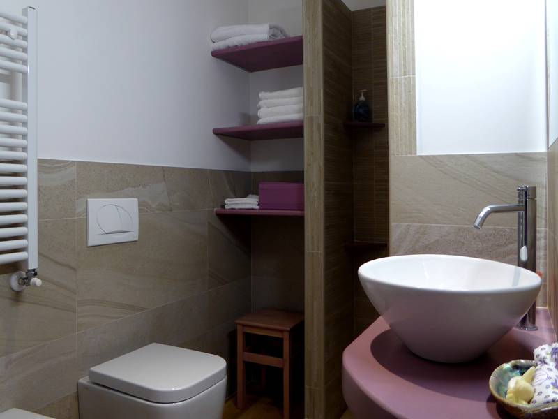 A modern bathroom is featured, showcasing a contemporary sink on a purple countertop. Shelves hold neatly arranged towels, while a compact stool is positioned beside the toilet. Natural light highlights the warm tones of the stone wall and offers a bright and clean environment.