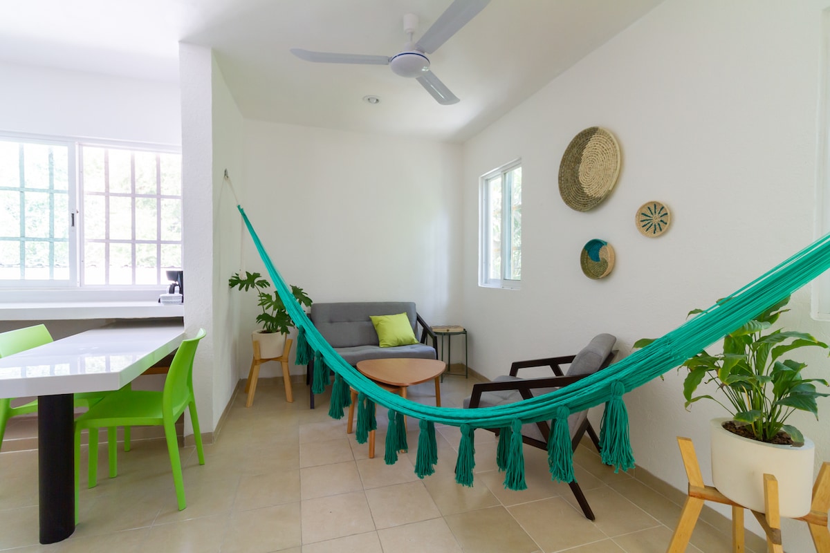 A bright and open living area features a spacious seating arrangement with a gray sofa and a light-colored coffee table. A colorful hammock is hung between two points, with decorative woven baskets on the wall. Large windows provide natural light, enhancing the neutral tones of the space.