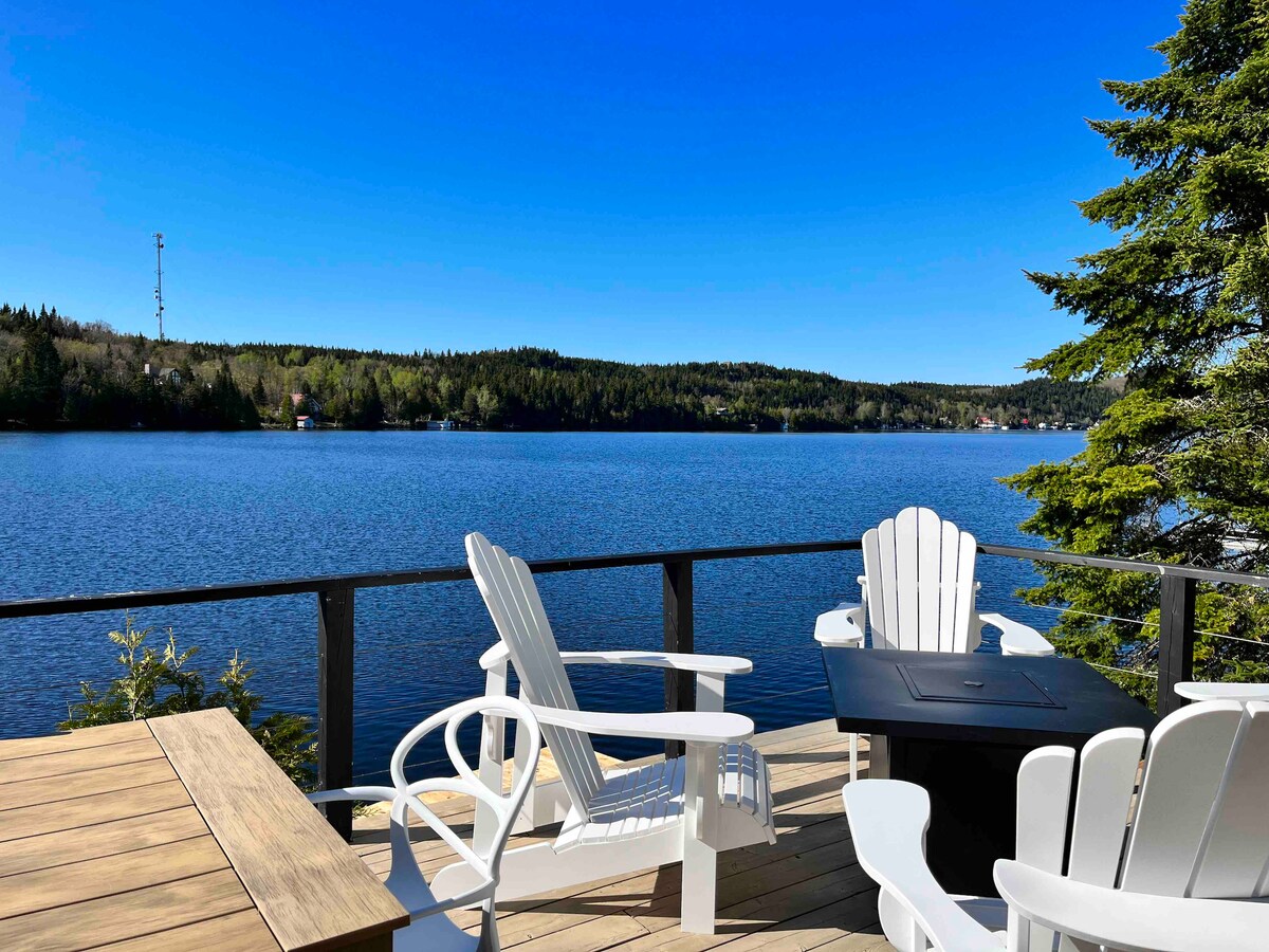 A spacious outdoor deck features white Adirondack chairs surrounding a fire pit, overlooking the serene waters of Lac Trois-Saumons. Lush green trees and clear blue skies enhance the tranquil lakeside setting, inviting relaxation and connection with nature.