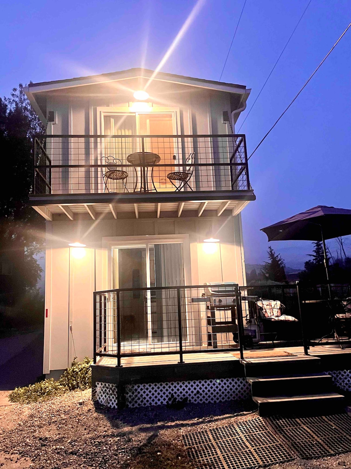 The two-story Carriage House is illuminated at dusk, showcasing a modern exterior. Two decks feature seating arrangements, with chairs and tables visibly arranged. The sliding glass doors invite a connection to the outdoors, while surrounding greenery adds a natural touch.
