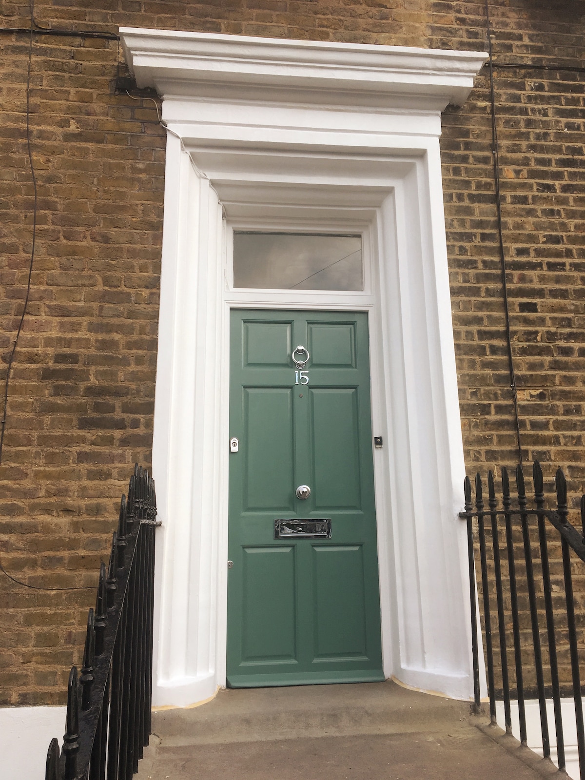 A traditional front door is seen, painted in a muted green shade. The door features multiple panels and a circular knocker, framed by a white, ornate architrave. A black wrought-iron railing runs along the path leading to the entrance.