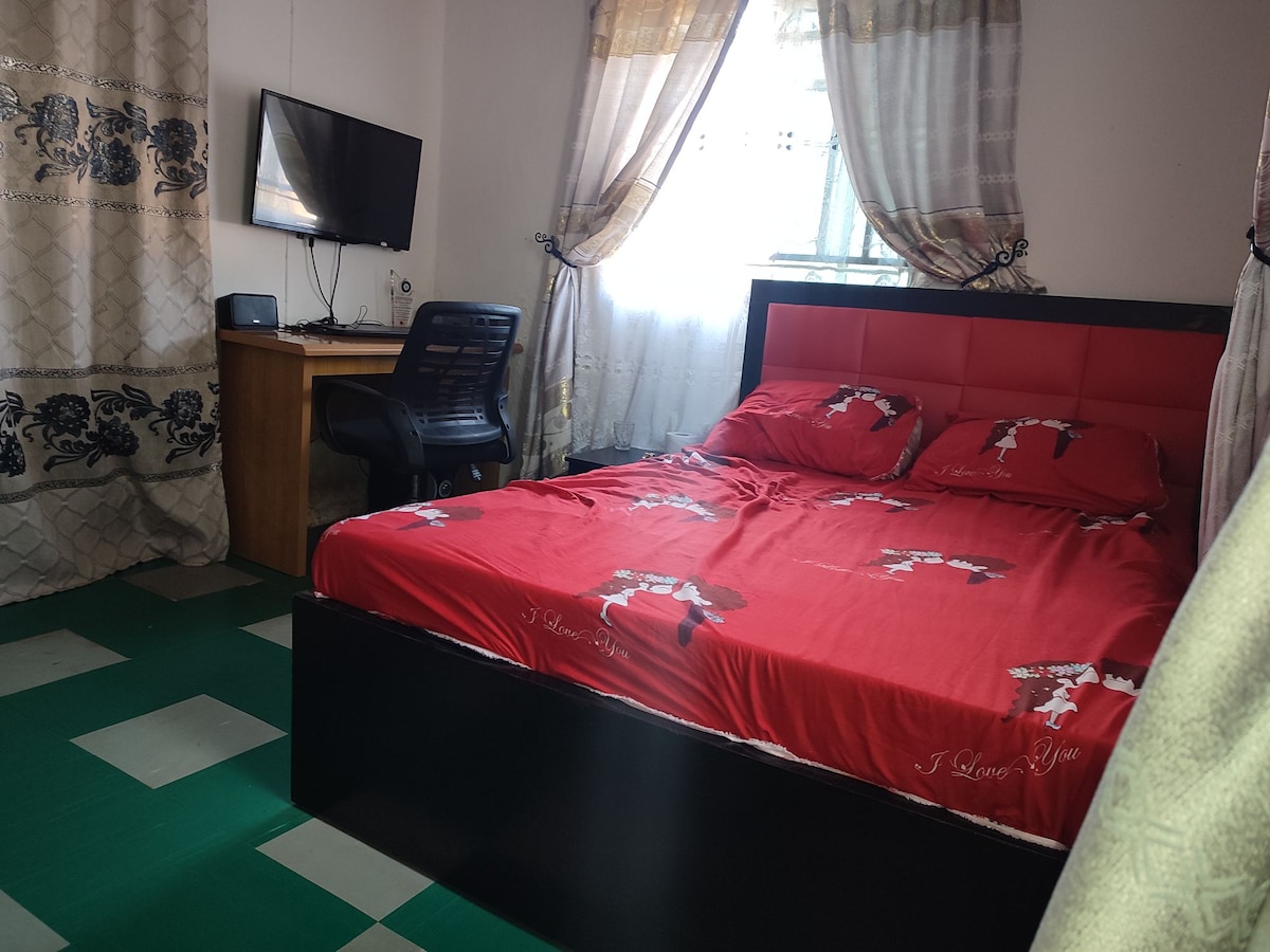 A cozy bedroom is equipped with a spacious bed dressed in red bedding adorned with simple motifs. A work desk and chair are positioned beside the window, where natural light filters through light curtains. A wall-mounted television is also visible.