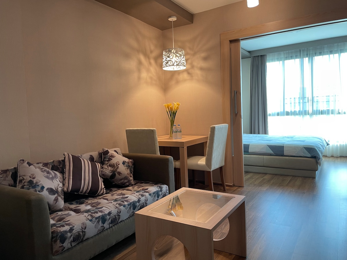 A cozy living area is depicted, featuring a floral-patterned sofa accompanied by a light wooden coffee table. A dining table for two is visible, with a vase holding yellow flowers. Natural light enters through large windows, illuminating the adjacent bedroom.