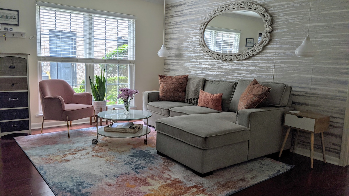 A light-filled living area features a spacious sectional sofa adorned with decorative pillows. A round glass coffee table sits on a colorful area rug. A cozy pink armchair and a reflective wall mirror enhance the room, while large windows provide views of greenery outside.