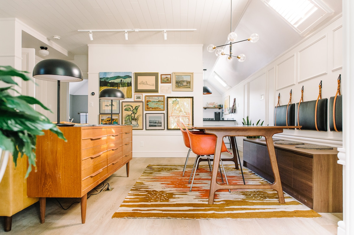 An inviting dining space is illuminated by natural light. A wooden table with orange chairs is centered on a colorful rug, with framed artwork adorning the walls. Stylish pendant lighting hangs above, complementing the modern aesthetic of the room.