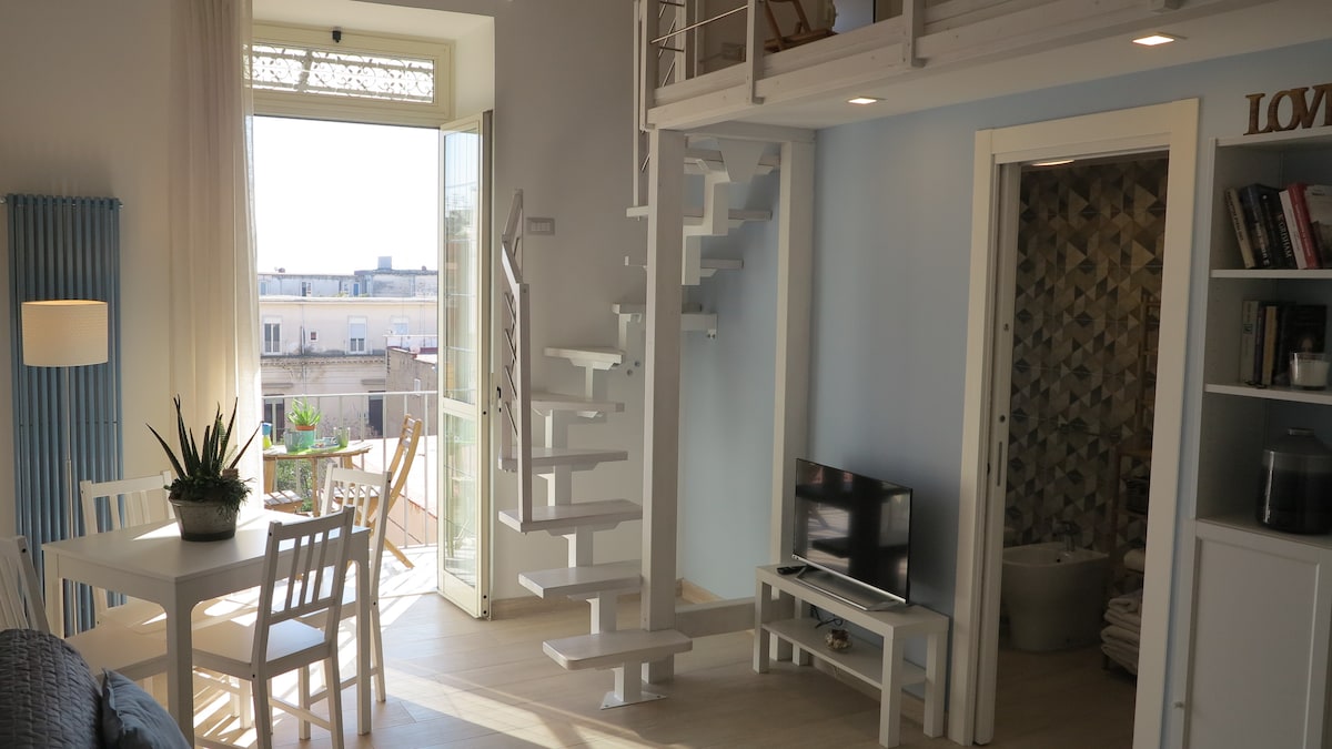 A bright living area features a white spiral staircase leading to a lofted sleeping space. A small TV sits on a white cabinet. A dining table with four chairs is positioned beside large open doors, offering views of a balcony adorned with greenery.