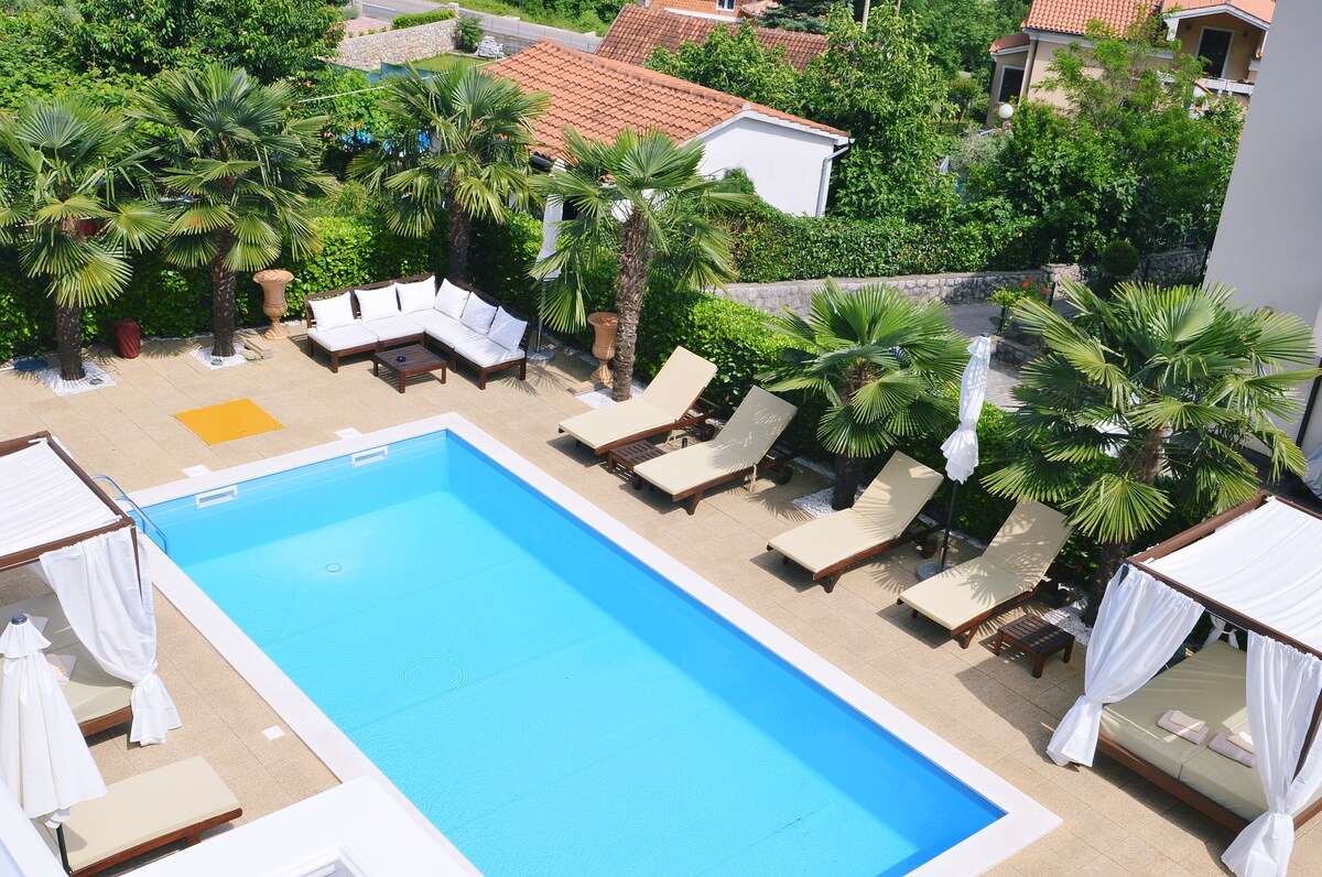 An outdoor pool area is surrounded by palm trees and sun loungers. Spacious poolside cabanas provide shaded seating. The clear blue water of the swimming pool reflects sunlight, offering a relaxing environment for guests. Lush greenery enhances the tranquil setting.