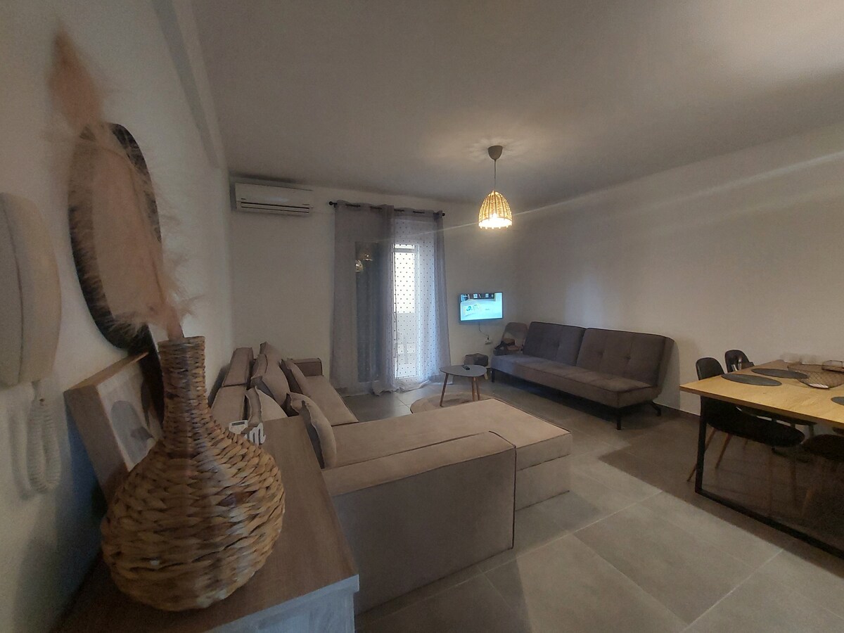 A spacious living area features two comfortable sofas arranged around a low coffee table. A television is mounted on the wall, while a dining table and chairs occupy one side of the room. Natural light enters through a door leading to a small balcony.