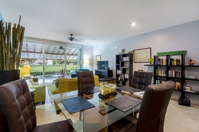 A modern living space features a glass dining table surrounded by dark chairs. A comfortable sofa in a vibrant green hue sits nearby. Large sliding glass doors open to the outdoor terrace, providing a view of the lush greenery beyond. A TV and bookshelf add functionality to the room.